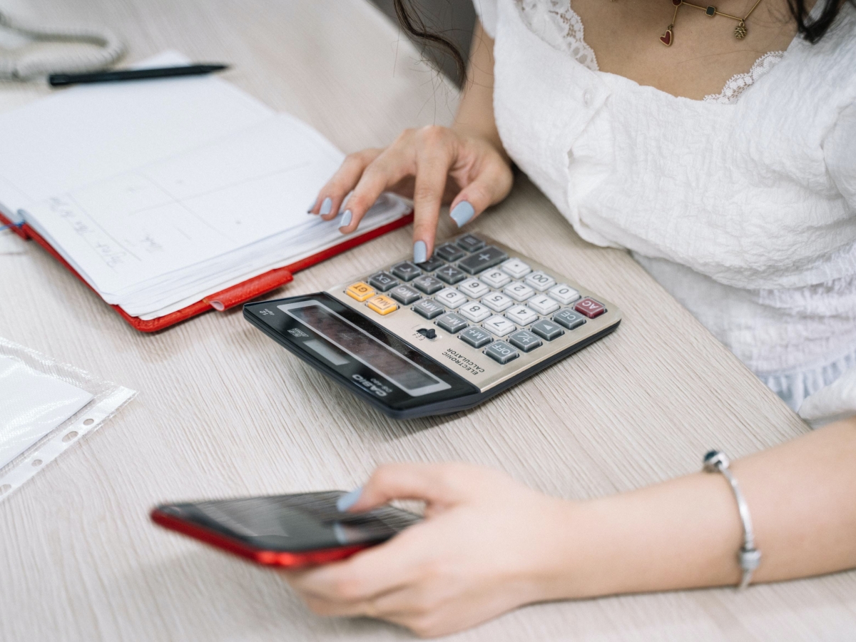LEAP a woman holding a red iphone, typing on a calculatore with a red notebook laying on the desk to her right.