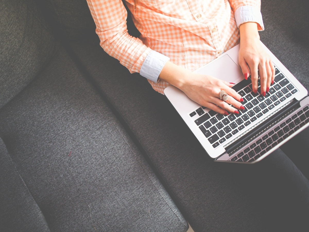 A person wearing an orange and white gingham patterned shirt works on a laptop while seated on a gray couch